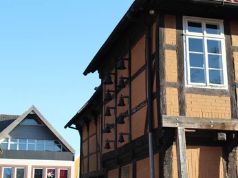Glockenspiel am Posthof Fachwerkhaus mit Glockenreihe an der Fassade, blauer Himmel, modernes Gebäude im Hintergrund.