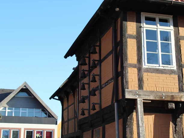 Glockenspiel am Posthof Fachwerkhaus mit Glockenreihe an der Fassade, blauer Himmel, modernes Gebäude im Hintergrund.