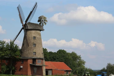 Pottmühle Petershagen Windmühle mit Flügeln und rotem Backsteingebäude, umgeben von Bäumen unter blauem Himmel.