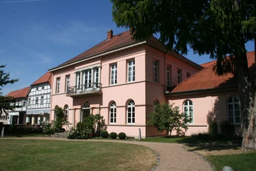 Historisches Herrenhaus in Pastellrosé mit großen Fenstern, umgeben von gepflegten Gartenanlagen.