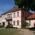Historisches Herrenhaus in Pastellrosé mit großen Fenstern, umgeben von gepflegten Gartenanlagen.