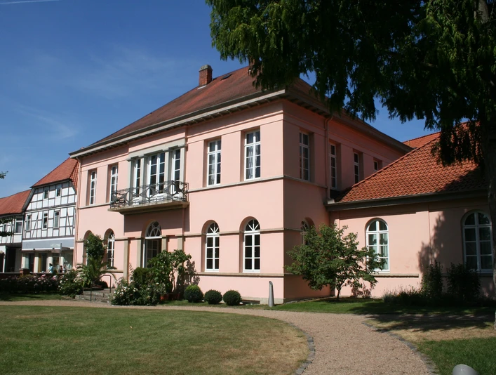 Historisches Herrenhaus in Pastellrosé mit großen Fenstern, umgeben von gepflegten Gartenanlagen.
