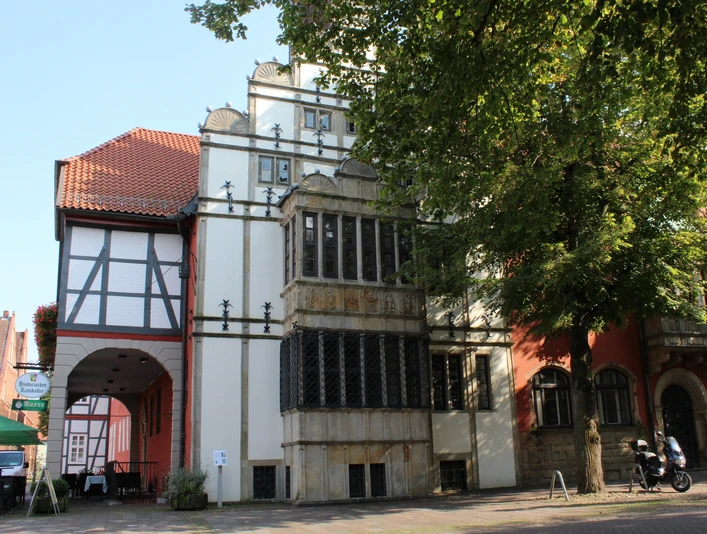 Rathausgiebel Historischer Rathausgiebel mit Fachwerkanbau bei Sonnenschein, flankiert von Bäumen und Bürgersteig.Historic town hall gable with half-timbered extension in the sunshine, flanked by trees and sidewalk.Historisk rådhusgavl med bindingsværkstilbygning i solskin, flankeret af træer og fortov.Historische gevel van het stadhuis met vakwerkuitbreiding in de zon, geflankeerd door bomen en bestrating.