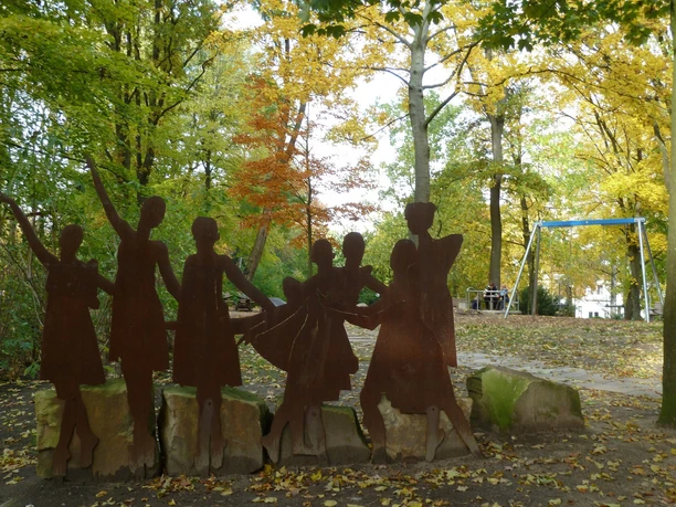Eine rostfarbene Stahlskulptur von tanzenden Menschen in einem herbstlichen Park mit bunten Blättern.
