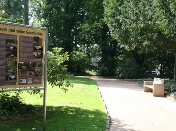 Ein geschwungener Weg führt durch den grünen, baumbestandenen Rathauspark in Achim, links ein Infoschild.