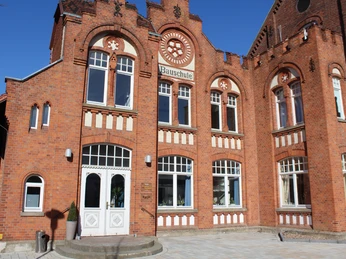 Historisches Backsteingebäude mit großen Fenstern, geschwungenen Verzierungen und einem zentralen Eingang.