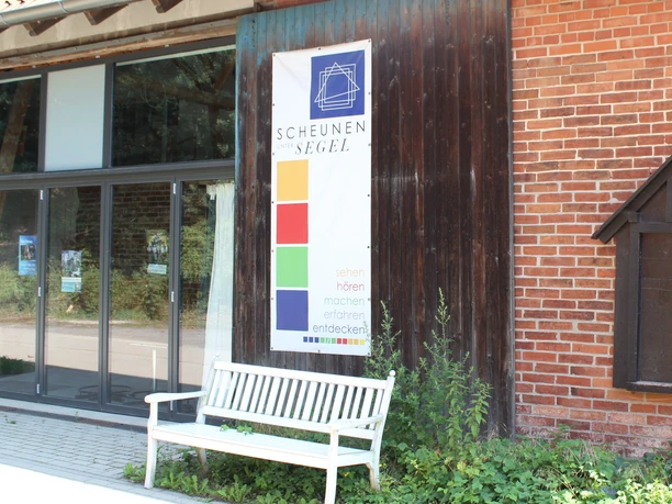 Außenansicht der Scheunenviertel Liebenau mit Info-Plakat, Bank und teils bewachsener Fassade.