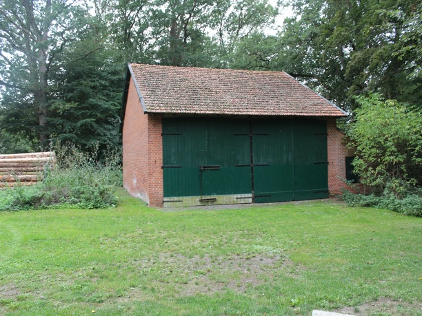 Scheunenviertel Wellie Eine rote Backsteinscheune mit grün gestrichenen Flügeltüren, umgeben von Bäumen und grünem Rasen.