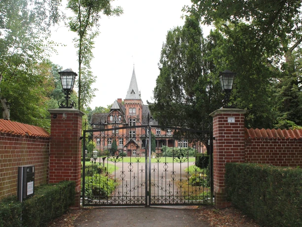 Schloss Eickhof Eine historische Backsteinvilla mit Turmspitze hinter einem schmiedeeisernen Tor und umgeben von Bäumen.
