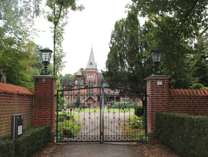 Schloss Eickhof Eine historische Backsteinvilla mit Turmspitze hinter einem schmiedeeisernen Tor und umgeben von Bäumen.