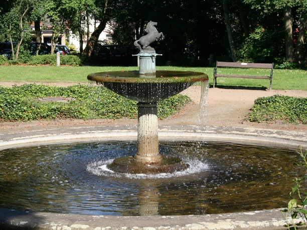 Fountain with horse statue, surrounded by green vegetation, trees and a park bench in the background.