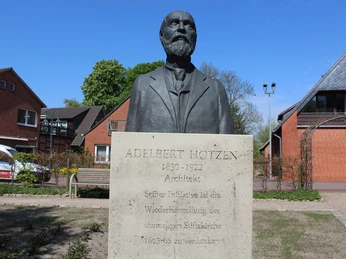 Skulptur eines älteren Mannes vor einem Sandsteinsockel mit Inschrift, umgeben von Backsteinhäusern.