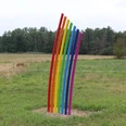 Eine bunte Skulptur aus gebogenen Streben in Regenbogenfarben steht auf einer grünen Wiese.