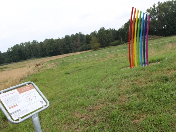 Skulptur "Am Ende des Regenbogens" Eine mehrfarbige Skulptur steht auf einer Wiese vor einem Wald. Ein Informationsschild ist sichtbar.