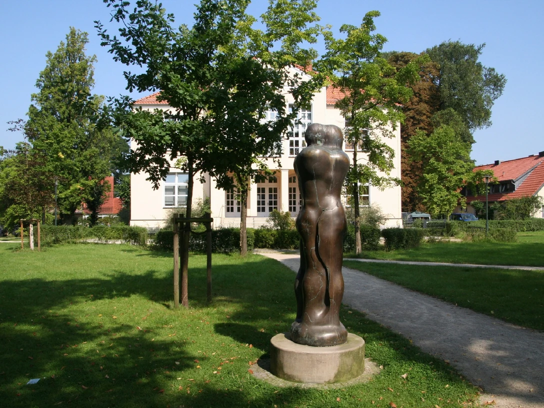 Skulptur Animus Anima steht im Park vor einem Gebäude, umgeben von Bäumen, im Sonnenschein.