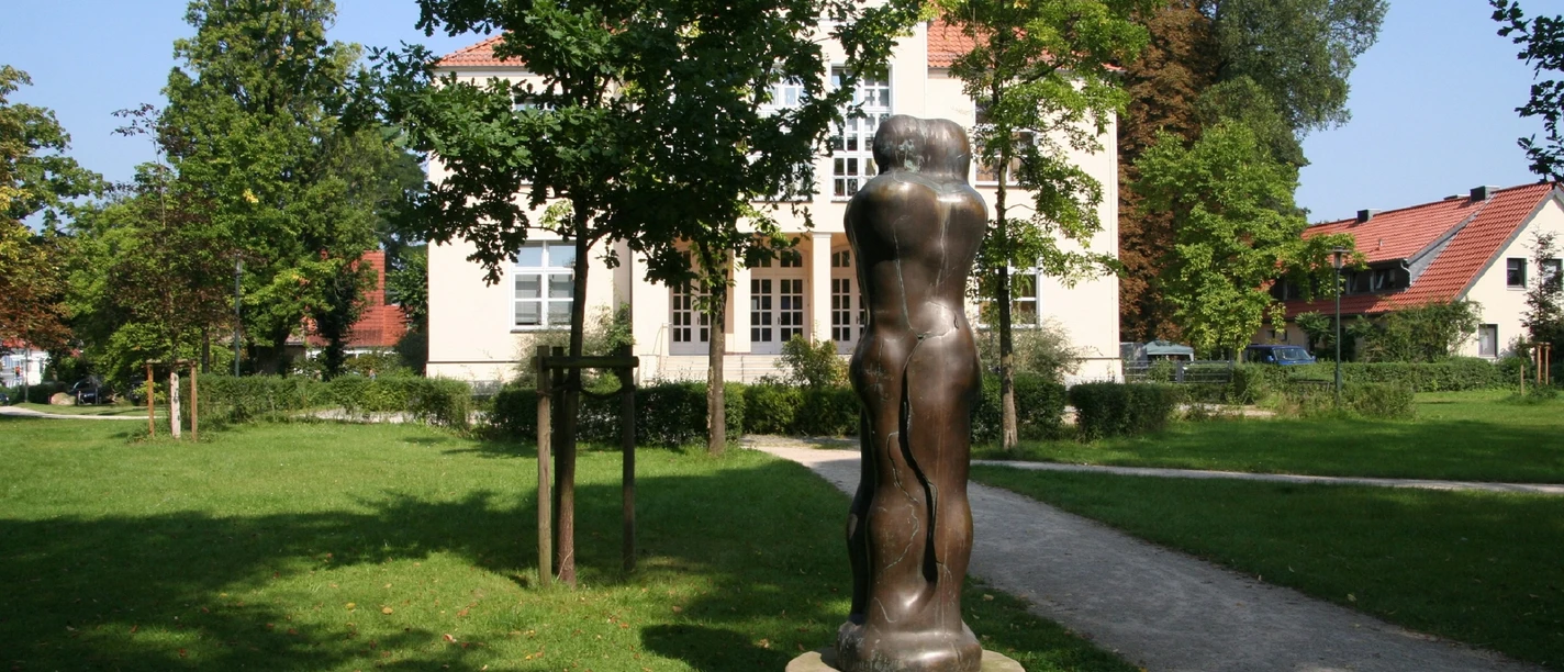 Skulptur Animus Anima steht im Park vor einem Gebäude, umgeben von Bäumen, im Sonnenschein.