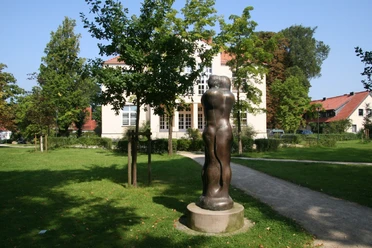 Skulptur Animus Anima Skulptur Animus Anima steht im Park vor einem Gebäude, umgeben von Bäumen, im Sonnenschein.