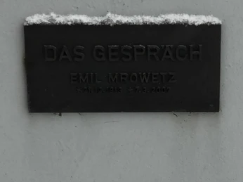 Schild an der Skulptur "Das Gespräch" Tafel mit der Inschrift "Das Gespräch" von Emil Mrowetz, beleuchtet durch Schneeakzente oben.