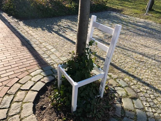 Ein weißer Stuhl ohne Sitzfläche umsäumt einen Baum, umgeben von Kopfsteinpflaster und Rasen.