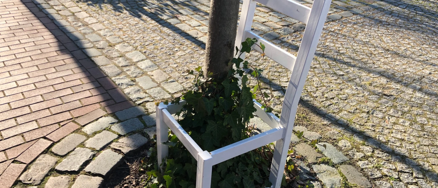 Ein weißer Stuhl ohne Sitzfläche umsäumt einen Baum, umgeben von Kopfsteinpflaster und Rasen.