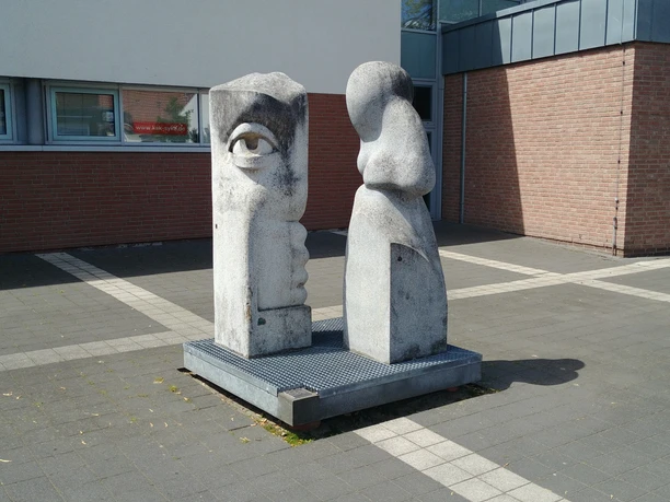 Skulptur aus zwei Steinfiguren mit Gesichtselementen vor einem modernen Backsteingebäude.