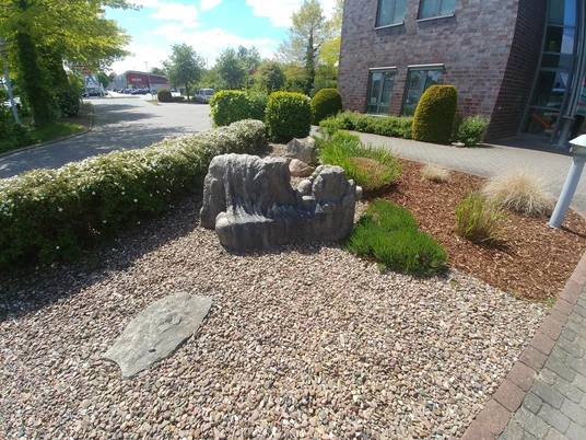 Steinskulptur einer liegenden Figur umgeben von Kiesbeet und grüner Bepflanzung vor einem Gebäude.
