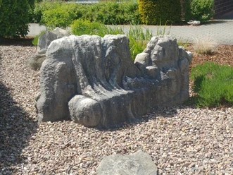 Steinskulptur mit liegenden Figuren und Gesicht auf einem Kiesbett, umgeben von Pflanzen und Gehweg.