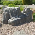 Die Liegende Steinskulptur mit liegenden Figuren und Gesicht auf einem Kiesbett, umgeben von Pflanzen und Gehweg.