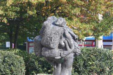 Eine Bronzeskulptur zeigt zwei sich umarmende Menschen, umgeben von üppigem Laubwerk.