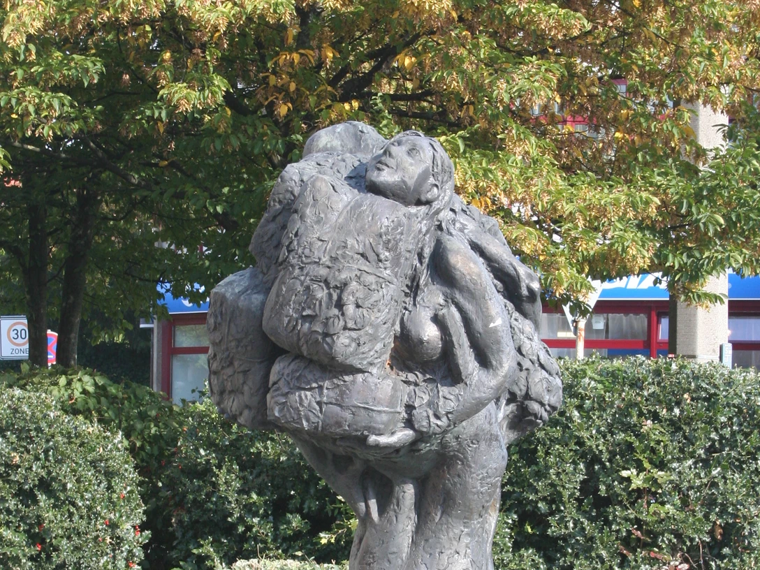 Eine Bronzeskulptur zeigt zwei sich umarmende Menschen, umgeben von üppigem Laubwerk.
