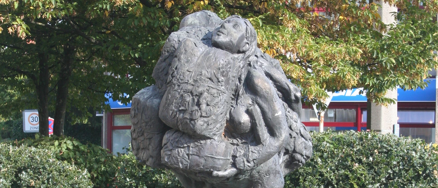 Eine Bronzeskulptur zeigt zwei sich umarmende Menschen, umgeben von üppigem Laubwerk.