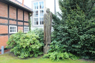 Eine stilvolle Engelsfigur aus Holz steht friedlich zwischen grünen Büschen vor einer Fachwerkfassade.