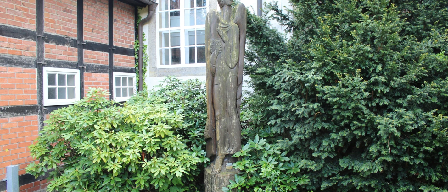 Eine stilvolle Engelsfigur aus Holz steht friedlich zwischen grünen Büschen vor einer Fachwerkfassade.