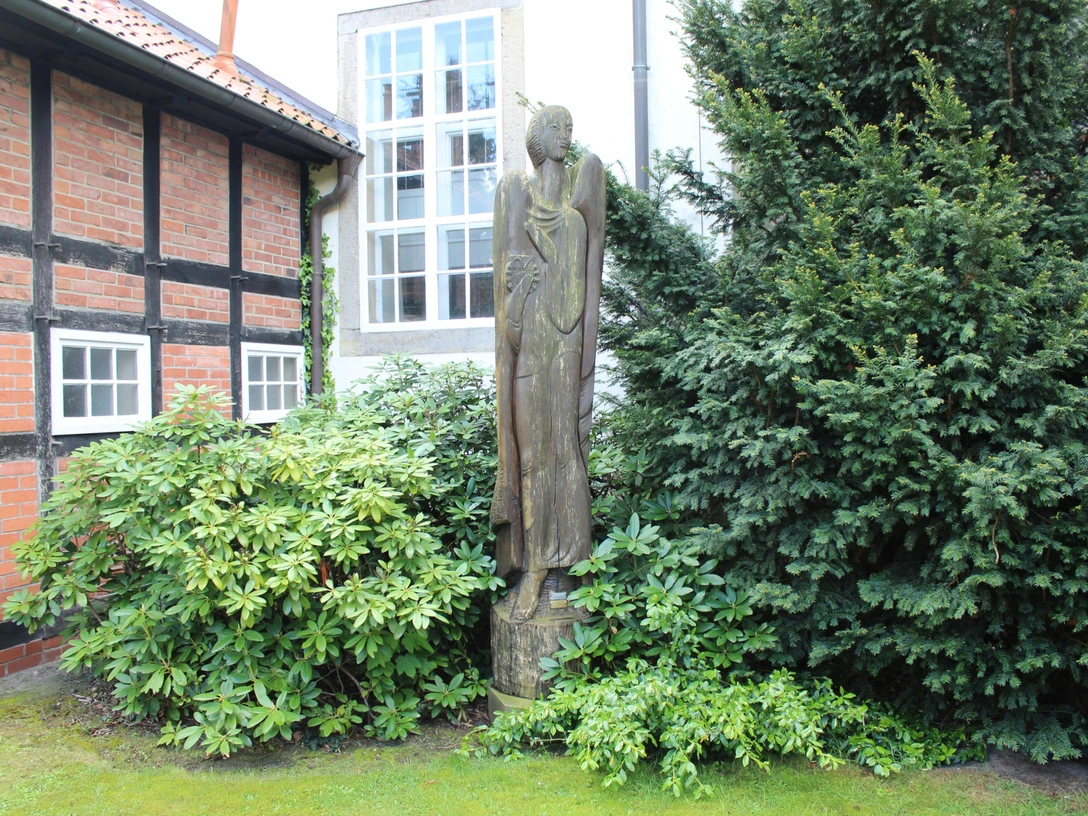 Skulptur Engel Warmsen Eine stilvolle Engelsfigur aus Holz steht friedlich zwischen grünen Büschen vor einer Fachwerkfassade.
