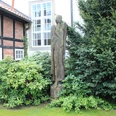 Skulptur Engel Warmsen Eine stilvolle Engelsfigur aus Holz steht friedlich zwischen grünen Büschen vor einer Fachwerkfassade.