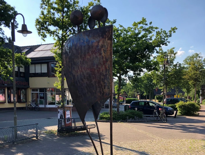 Zwei abstrahierte Figuren in moderner Metallskulptur stehen vor einer Apotheke im städtischen Ambiente.