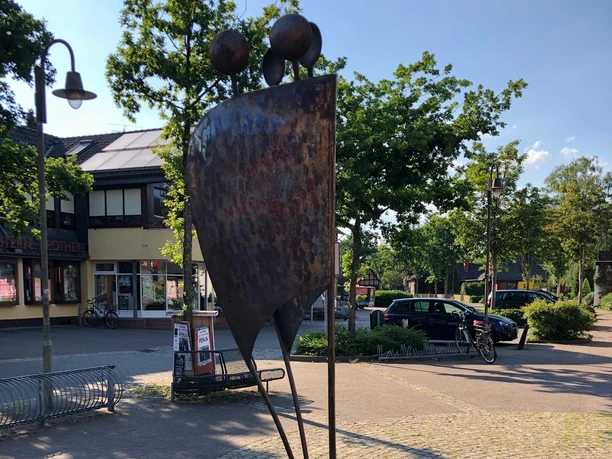 Skulptur Entfaltung Zwei abstrahierte Figuren in moderner Metallskulptur stehen vor einer Apotheke im städtischen Ambiente.