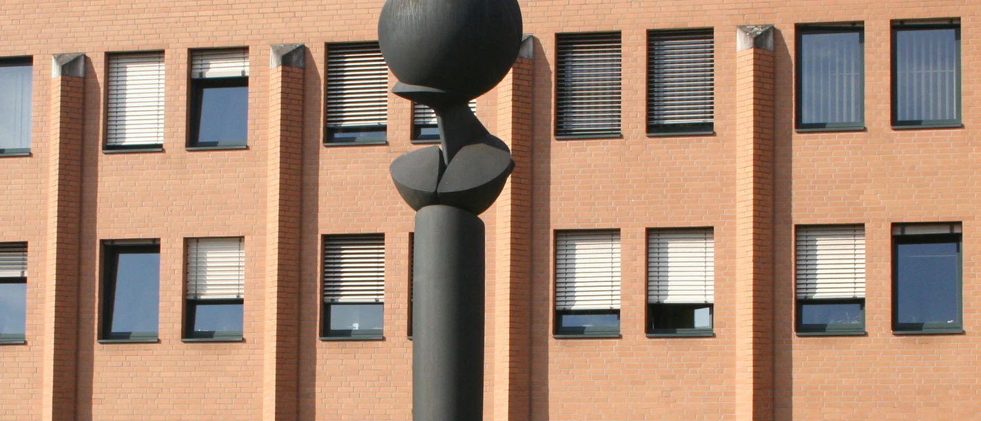 Skulptur mit kugelförmiger Spitze vor einem Backsteingebäude mit zahlreichen quadratischen Fenstern.
