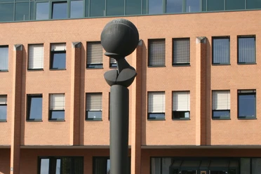Flora II Skulptur mit kugelförmiger Spitze vor einem Backsteingebäude mit zahlreichen quadratischen Fenstern.