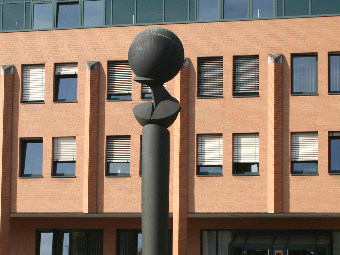 Flora II Skulptur mit kugelförmiger Spitze vor einem Backsteingebäude mit zahlreichen quadratischen Fenstern.