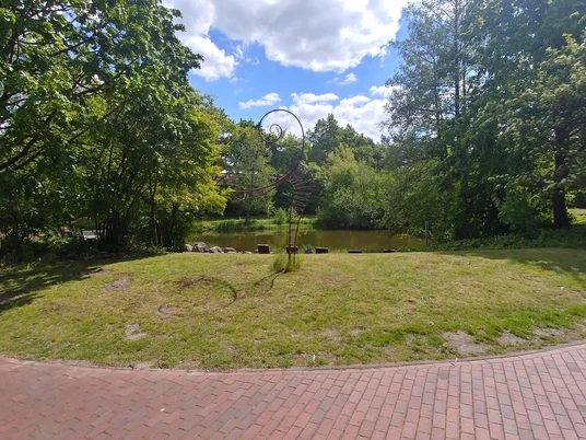 Füllhorn oder Figura Metallskulptur auf einer grünen Wiese vor einem Teich, umgeben von Bäumen. Himmel leicht bewölkt.