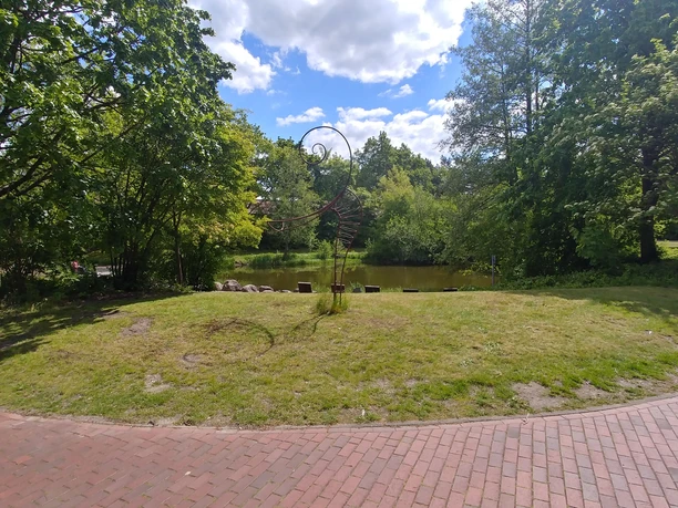 Füllhorn oder Figura Metallskulptur auf einer grünen Wiese vor einem Teich, umgeben von Bäumen. Himmel leicht bewölkt.
