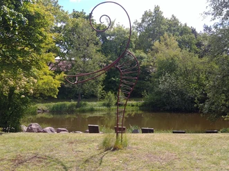 Eine spiralige Metallskulptur steht vor einem Teich in einem parkähnlichen Bereich, umgeben von Bäumen.