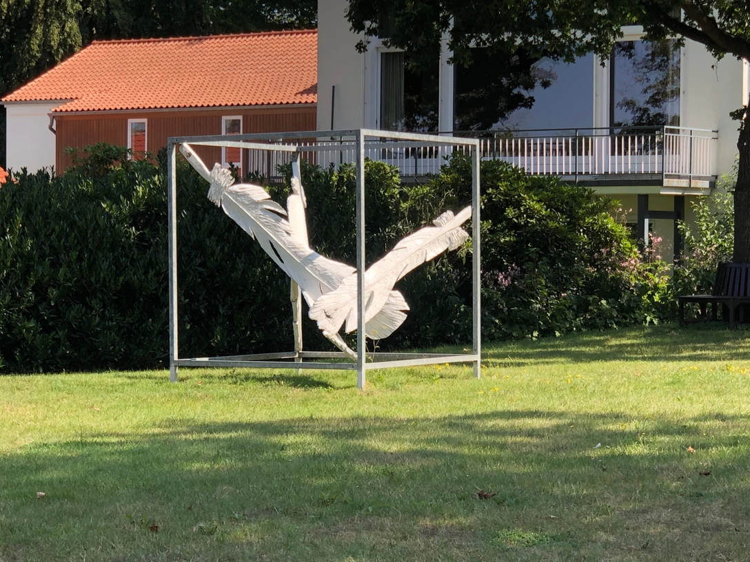 Gefallen Eine skulpturale Darstellung von verknoteten weißen Stoffstreifen in einem quadratischen Metallrahmen auf einer grünen Rasenfläche. Im Hintergrund sind dichtes Gebüsch und ein modernes Gebäude mit rotem Ziegeldach und Balkon zu sehen.