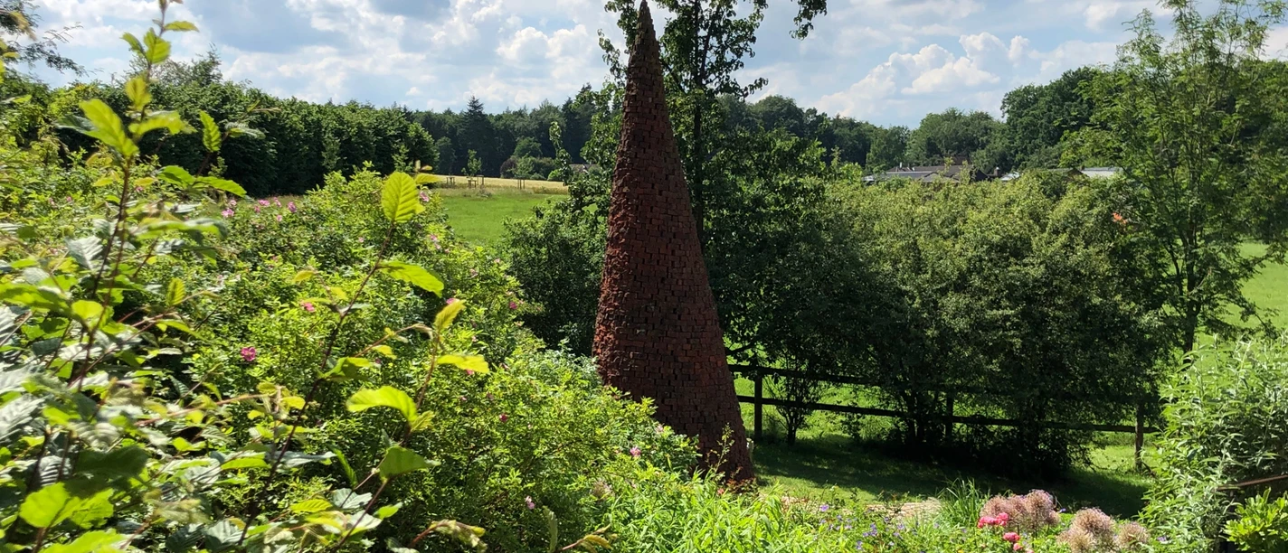 Ein markanter, spitzer Ziegelturm erhebt sich inmitten einer grünen und bewachsenen Landschaft.