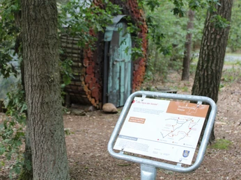 Skulptur "Haus der Sonne" Holzskulptur in einem bewaldeten Gebiet, umgeben von Bäumen, mit einer Informationskarte im Vordergrund.