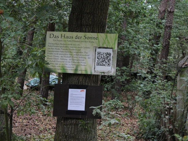 Ein Schild im Wald informiert über die Skulptur "Haus der Sonne" mit QR-Code, umgeben von Bäumen.