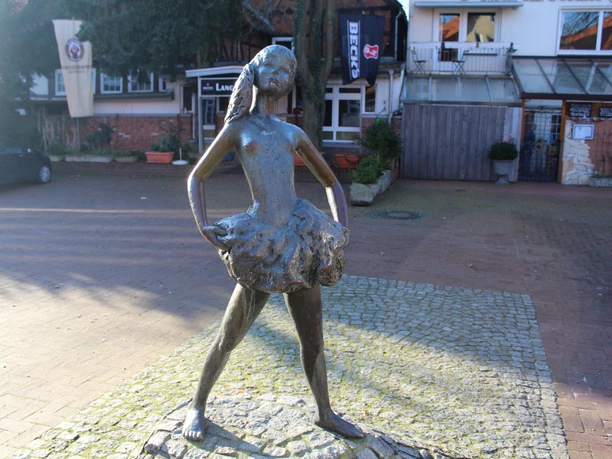Bronzestatue eines jungen Mädchens in Ballerinapose auf gepflastertem Platz vor Geschäften.