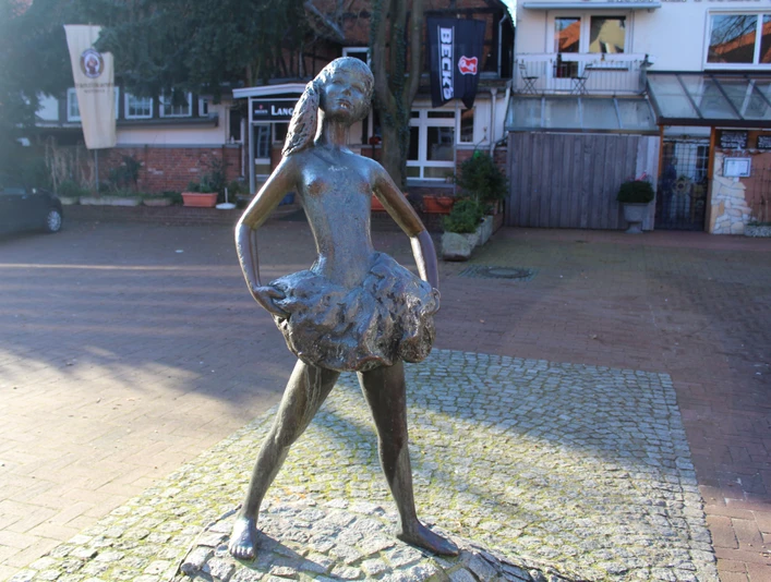 Bronzestatue eines jungen Mädchens in Ballerinapose auf gepflastertem Platz vor Geschäften.
