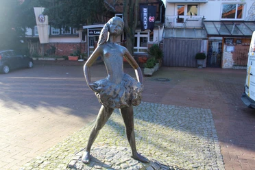 Kleine Nienburgerin Bronzestatue eines jungen Mädchens in Ballerinapose auf gepflastertem Platz vor Geschäften.